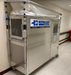 HEPACART 74" / 55" AutoLift Mobile Containment Unit with Built-in Air Scrubber tent attached to the top. The unit is on wheels and has a door on the right side. It also has a control panel and viewing window on the front.  The HEPAcart logo is visible on the side.  It appears to be set up in a hallway, possibly in a hospital or laboratory setting.